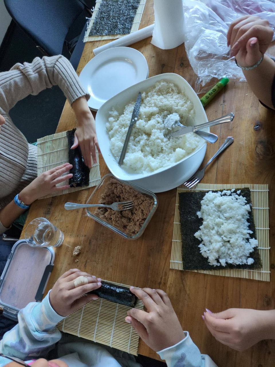 Projektbild am Küchentisch. Zu sehen ist ein Tisch von oben auf dem gerade Sushi zubereitet wird. Drei Händepaare rollen Sushi oder greifen in die Reisschüssel. Die Jugendlichen tragen teilweise Armbänder oder Ringe an den Händen.